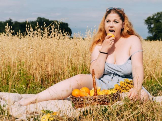 Jessika Daniels mit orangefarbenen Brüsten, die eine Pfirsich isst, auf einem Feld mit einem Korbkorb voller Orangen und gelber Blumen, sitzend auf einer Decke, trägt ein blaues Kleid..