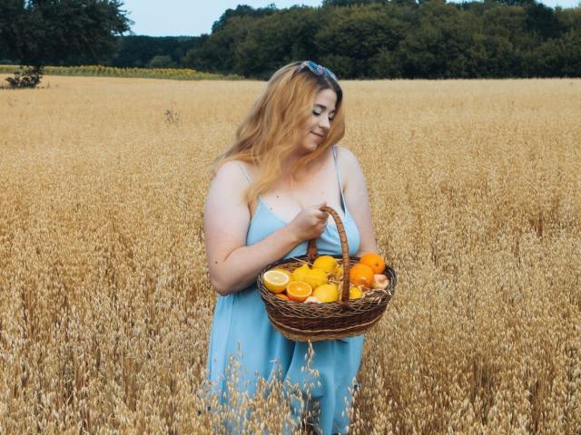 Jessika Daniels mit welligem, blondem Haar, trägt ein blaues Kleid und hält einen Korb mit Orangen und geschnittenen Orangen. Sie steht in einem goldenen Weizenfeld. Ihre Brüste sind sichtbar..