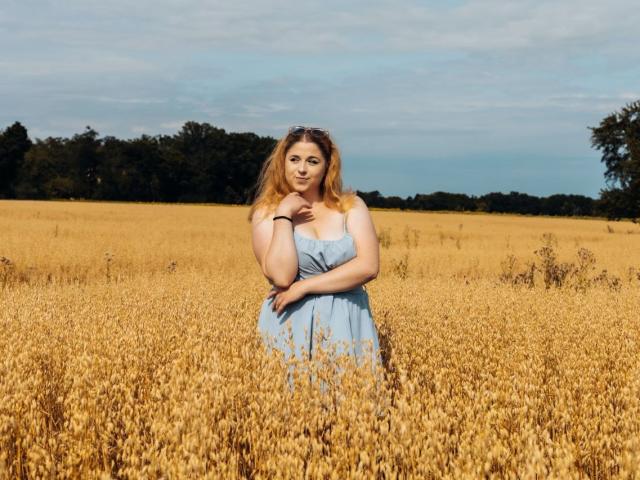 Jessika Daniels steht in einem goldenen Weizenfeld, trägt ein blaues, trägerloses Kleid und hält ihre Hand in die Nähe ihres Kinns. Sie schaut in die Kamera, wobei ihr Hintern leicht sichtbar ist..