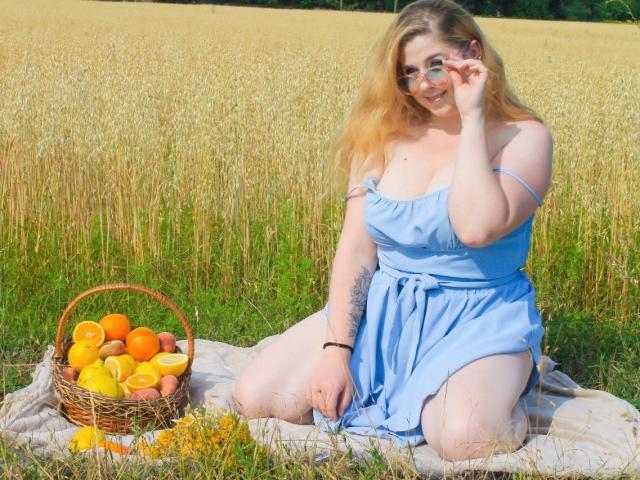 Jessika Daniels, rubia y con gafas, arrodillada en un campo, vestida con un vestido azul, sosteniendo unas gafas, con una cesta de mimbre llena de naranjas y melocotones sobre una manta..