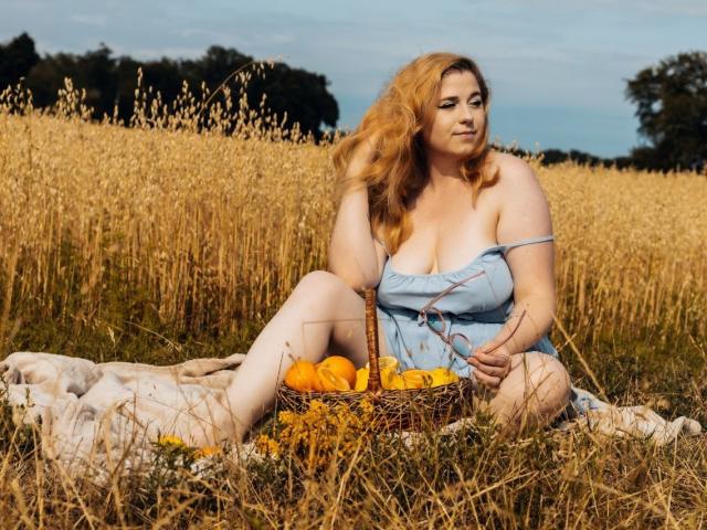 Jessika Daniels, con grandes senos y piel pálida, está sentada en un campo, vestida con un vestido azul, sosteniendo una cesta de naranjas, y tiene una manta debajo, con flores amarillas..
