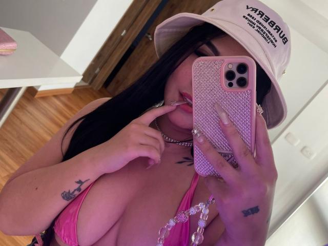 SweetEmily69 with long black hair, pink bikini showing big tits, white bucket hat, pink phone case, finger in mouth, tattoos on hand and chest, wooden floor.