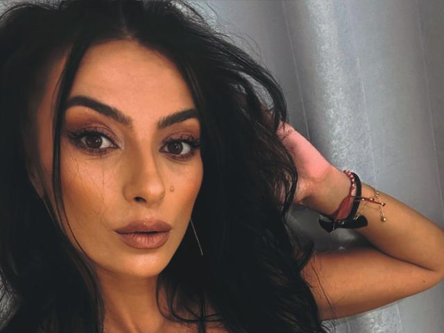 KinkyAmelie has dark, wavy hair, thick eyebrows, brown eyes, and a small beauty mark on her left cheek. She's wearing subtle makeup and a bracelet on her right wrist.