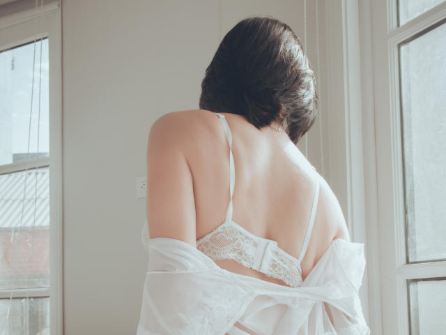 LatinLola with short brown hair, facing away, wearing white lace bra, sheer white shirt off her shoulders, sunlight through window, soft natural light, visible back, subtle elegance.
