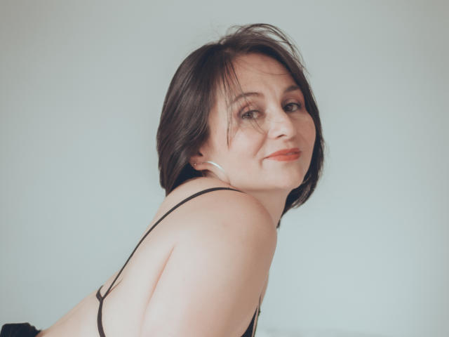 LatinLola has short brown hair, fair skin, and a slight smile. She's wearing a black strap top, showing her bare shoulders and back. She's looking over her shoulder.