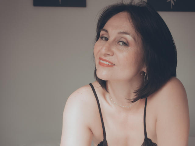 LatinLola with short black hair, smiling at the camera, wearing a black spaghetti strap top, showing her bare shoulders and a delicate necklace. She's got cute tits and a subtle smile.