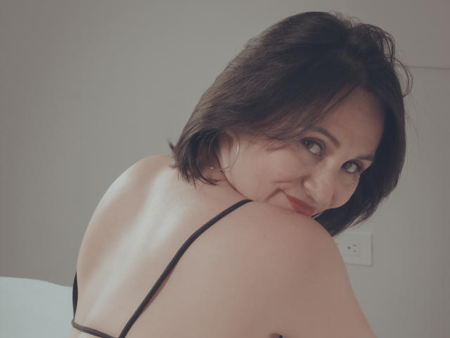 LatinLola with short dark brown hair, smiling at the camera, showing her bare back and black strap, looking over her shoulder, soft light, slight red lipstick.
