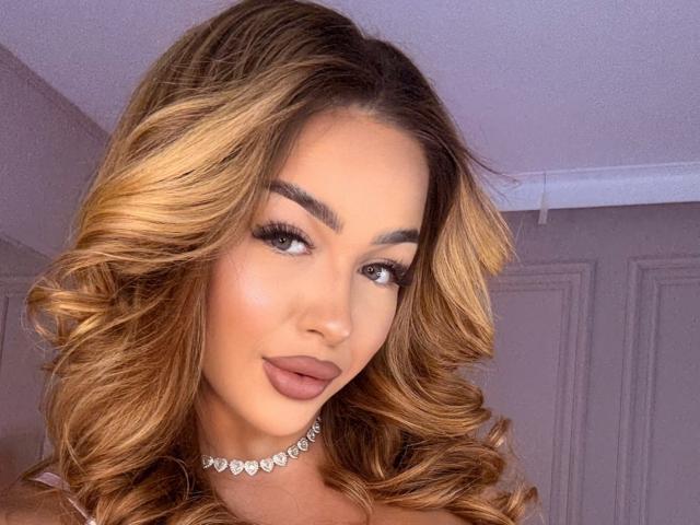 xJuliax has curly blonde hair, full eyebrows, long lashes, pink lipstick, and a heart-shaped choker, looking seductive with a slight smile.