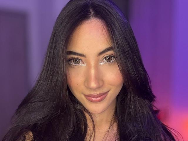 JuanaFox has long black hair, brown eyes with glittery makeup, and a cute smile. Her pink lips are slightly parted. She's looking directly at the camera.