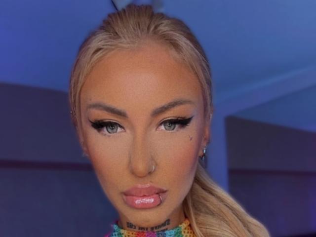 SinnersMuse has blonde hair, green eyes, and full pink lips with a small nose ring. Her makeup is dramatic with thick eyeliner and a subtle tattoo below her chin.