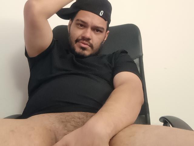StivensAndWilliams is a chubby, bearded man with light brown skin, wearing a black cap and shirt, sitting with his legs spread, exposing his hairy, uncircumcised, flaccid penis.