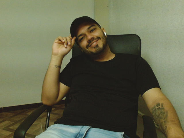 StivensAndWilliams, a chubby dude with a black cap, beard, and tattoo on his forearm, wearing a black tee and light blue jeans, sitting in a chair, smiling.