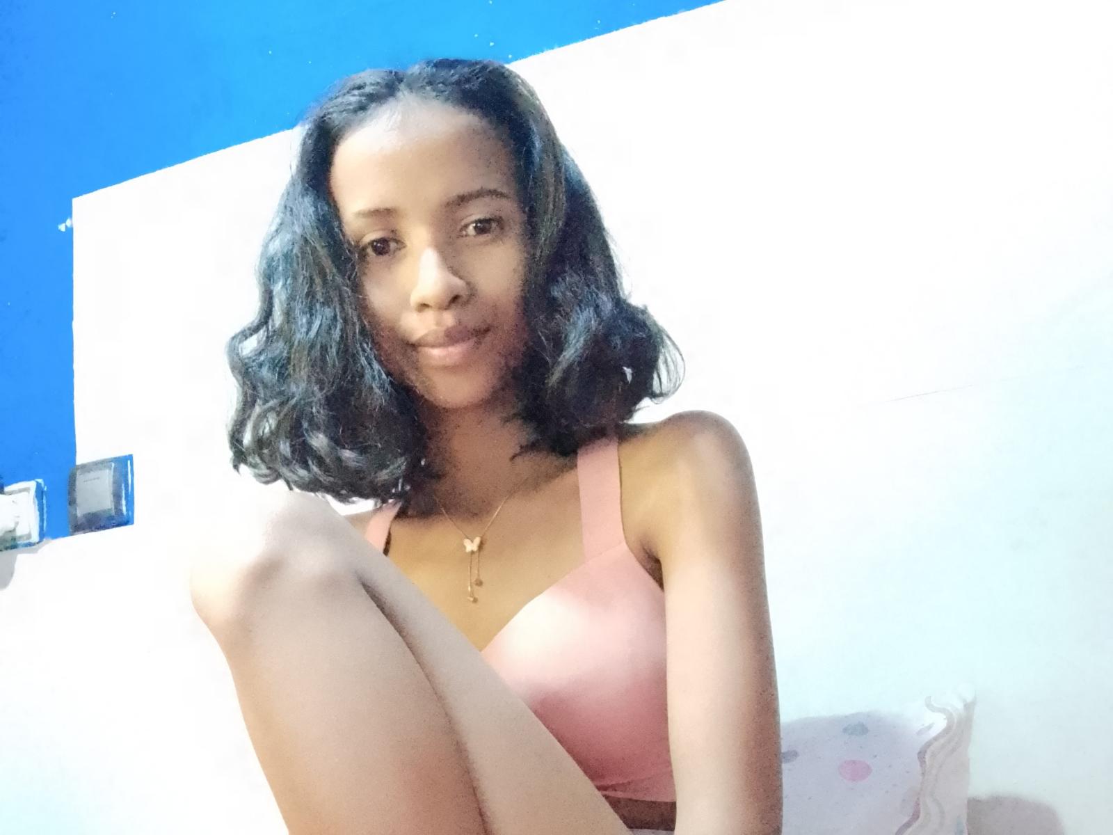 JohanneX has medium brown skin, black wavy hair, small tits in a pink bra, sits with knees up, smiling, wearing a gold necklace, in a brightly lit room.