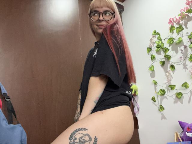 KallySpice with long red and blonde hair, glasses, black shirt, and no pants, showing her tattooed thigh and smiling at the camera, her ass slightly raised.