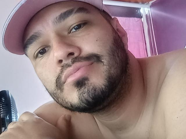 StivensAndWilliams has a light brown beard, dark eyebrows, and a pink cap, with a slight smile, shirtless, holding a black hair dryer, and has a medium skin tone.