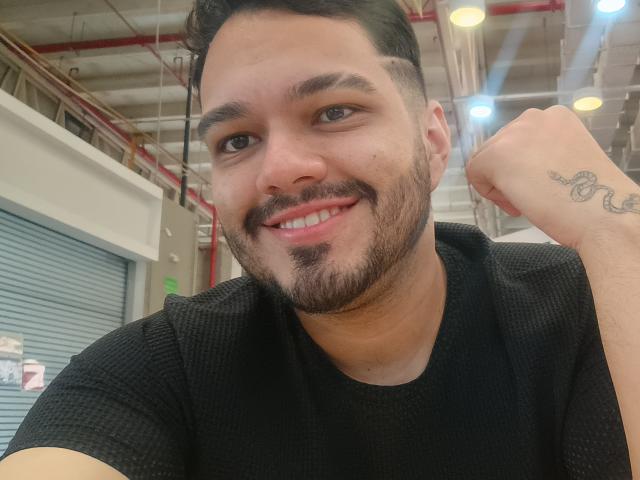 StivensAndWilliams with a smiling face, short black hair, beard, tattoo on right forearm, black waffle-texture shirt, raising right fist, brown eyes, and light skin.