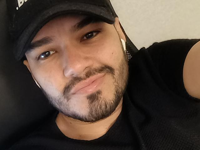 StivensAndWilliams with a light brown skin tone, black baseball cap, small goatee, black t-shirt, white earbuds, slightly smiling, brown eyes, short black hair, and a relaxed expression.