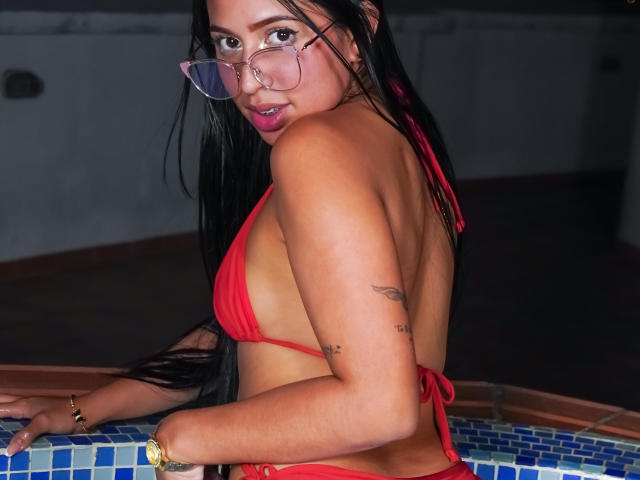 Ciamsth with long black hair, wearing a red bikini, glasses, and gold watch, looking over her shoulder, showing her tattoos and small boobs, standing by a tiled pool.