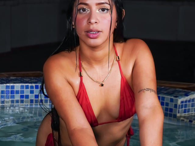 Ciamsth has wet, dark hair, red bikini, gold necklace, and tattoo on right arm, leaning in a pool, showing her tits and small ass, with water on her skin.