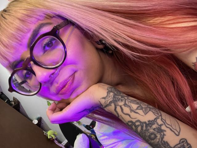 KallySpice with pink and orange hair, large round glasses, and a tattooed arm, resting her chin on her hand, showing a cute smile with purple lighting.