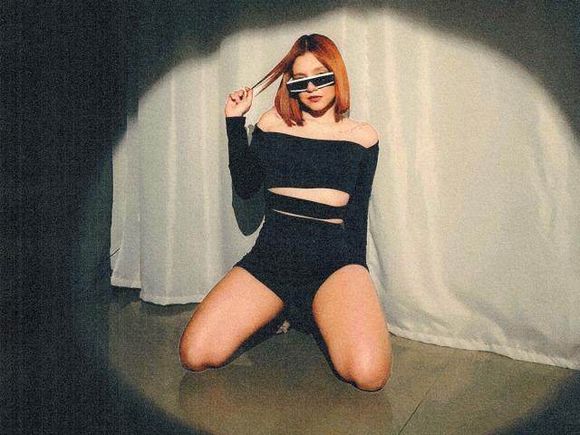 AmbeerLux, kneeling, wearing black off-shoulder top with cutouts, black shorts, and white sunglasses, holding hair, with visible ass, in dimly lit room, shadow on wall.