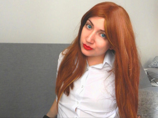 BambiSoranjo-hot a de longs cheveux roux, des yeux bleus et du rouge à lèvres. Elle porte une chemise blanche à boutons et est assise sur un canapé gris avec un coussin moelleux..