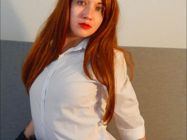BambiSoranjo, une femme aux longs cheveux roux, portant une chemise blanche à boutons, laissant entrevoir légèrement ses seins à travers le tissu, avec des lèvres rouges, des yeux bleus et un sourire charmant..
