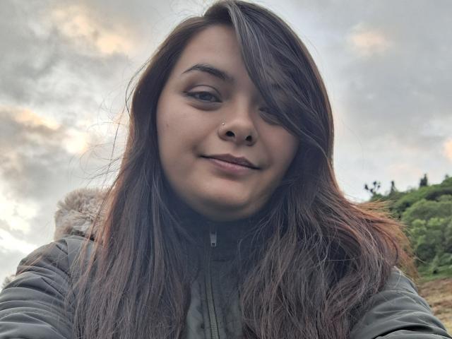 KatherinLatin has long brown hair, a nose ring, and a slight smile, wearing a green jacket with a furry hood. Her small tits are slightly visible under the jacket.