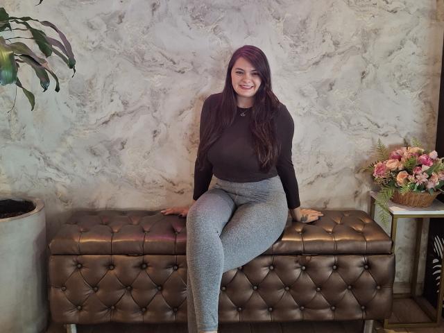 KatherinLatin with long brown hair, black top, gray leggings, sitting on brown tufted bench, smiling, hands on bench, pink flowers on right, green plant on left.