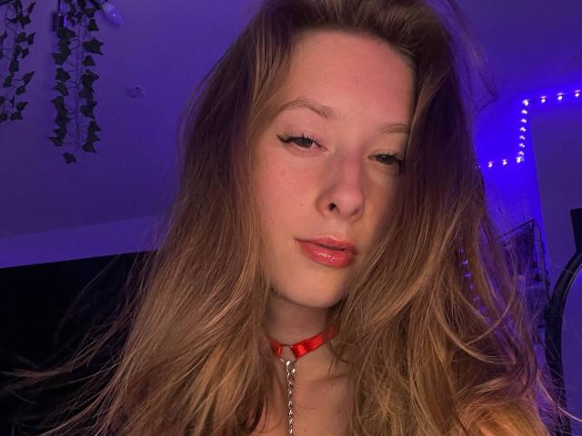 CandyFlosse with long brown hair, red collar, and chain, winking with glossy pink lips, in a purple-lit room. Her face is slightly flushed with a cute, sexy look.