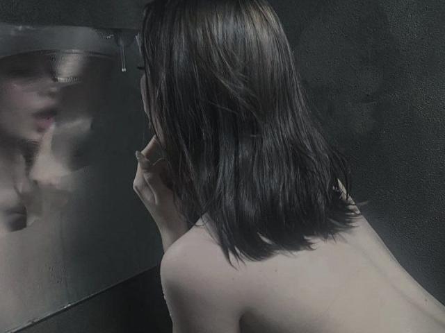 LollaKiss with shoulder-length brown hair, naked, gazing at a fogged-up mirror. Her reflection shows a blurred, partially visible face. Her hand touches the mirror.
