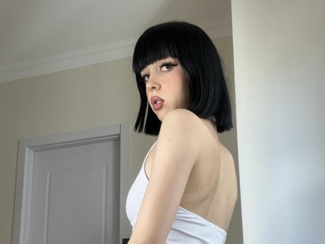 SaraMiner with black bob, white tank top, showing her back and small tits, looking over her shoulder, tongue out, in a room with a white door.