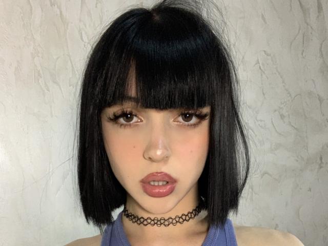 SaraMiner with black bob, brown eyes, thick eyelashes, pink lips, choker, and purple tank top, showing a neutral expression. Her small nose and smooth skin are visible.