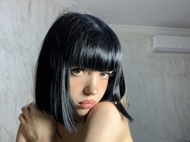 SaraMiner with straight black bob, brown eyes, and shiny lips, arms crossed over small bare breasts, looking up with serious expression, in a softly lit room.