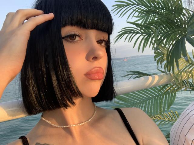 SaraMiner with black bob, hand in hair, pearl necklace, brown eyes, glossy lips, black straps, palm leaves, and ocean in background, sunlight highlighting her face.