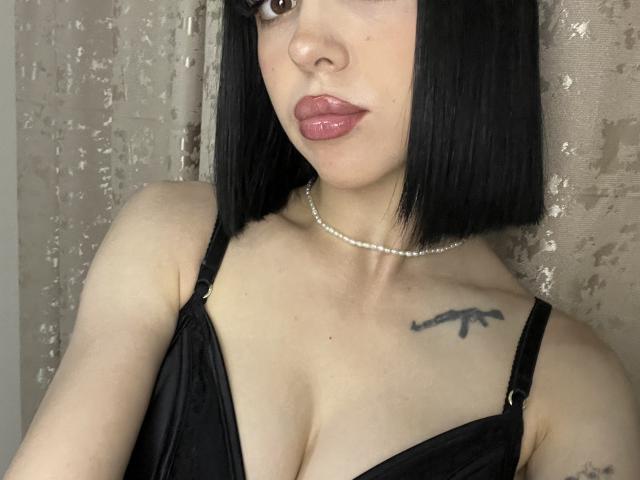 SaraMiner with black bob, pink lips, pearl necklace, black dress, showing her tits, tattoo on right shoulder, fair skin, looking at camera.