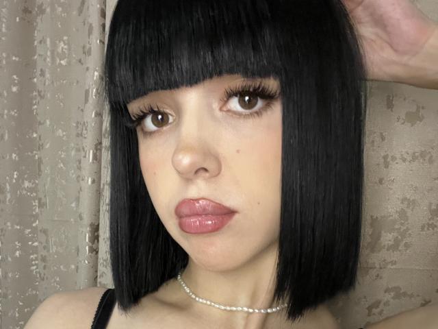 SaraMiner has a sleek black bob with bangs, brown eyes, glossy pink lips, and a pearl necklace. She's wearing a black top, with a hand in her hair.