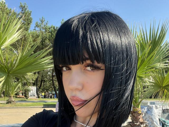 SaraMiner with glossy black straight hair, bangs, and long eyelashes, pouting with pink lips, wearing a black top and pearl necklace, standing in front of green palm trees.
