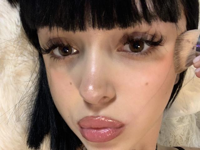 SaraMiner's face, glossy pink lips, dark brown eyes, long black eyelashes, straight black hair with bangs, light skin, small moles, and shiny nose. She's holding her right ear.