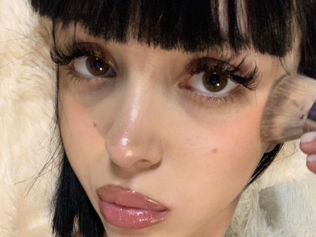 SaraMiner has glossy pink lips, dark brown eyes with long eyelashes, and a black-haired face with blunt bangs, while holding a brush against her right cheek.