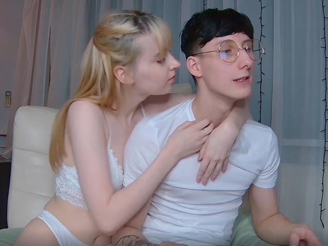 Blonde VipCouples with small tits and white lace bra kisses dark-haired VipCouples with glasses and white tee, arm around neck, in cozy room.