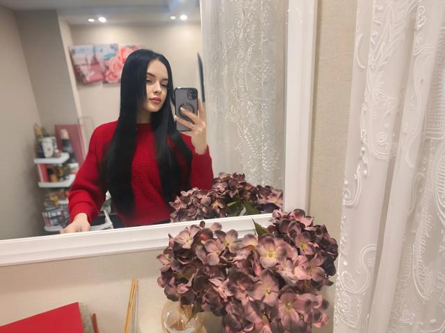 Lulii69 with long black hair, red sweater, taking mirror selfie, holding phone, standing in front of white-framed mirror with purple flowers in foreground.