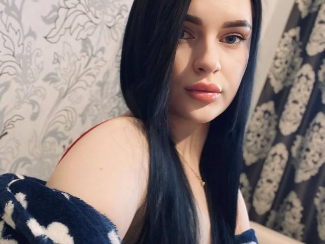 Lulii69 with long black hair, fair skin, and pink lipstick, wearing a navy blue off-shoulder top, showing her shoulder and subtle nose ring, looking at the camera.