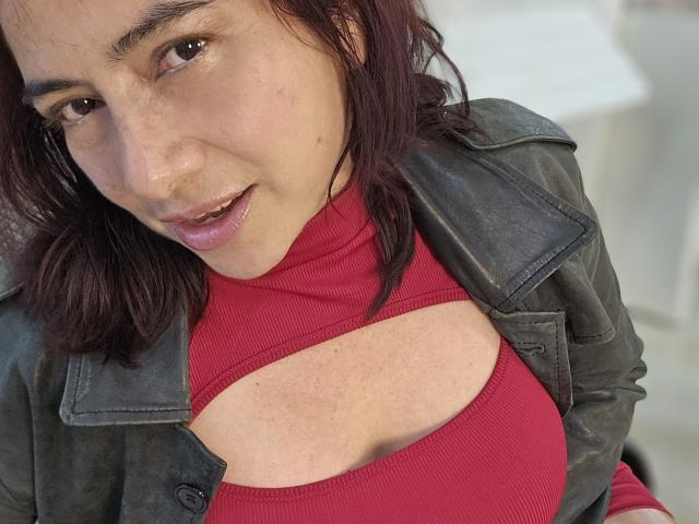 Alessia: Hellhäutige Frau mit mittlerem Hautton, dunkelrotem Haar und einem leichten Lächeln, die ein rotes, geripptes Oberteil mit Ausschnitt und eine grüne Lederjacke trägt. Brüste sind sichtbar..