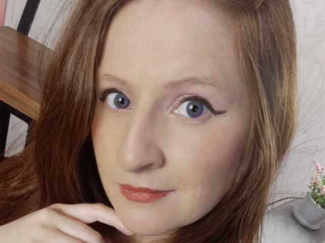 EnzumGinger's face, showing blue eyes with black eyeliner, red lips, and light brown hair. Her hand rests near her chin, with a subtle, confident expression.