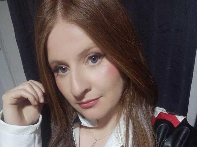 EnzumGinger has light brown hair, blue eyes, pink cheeks, and a slight smile. She's wearing a white shirt with a black and red jacket. Her hand is near her face.