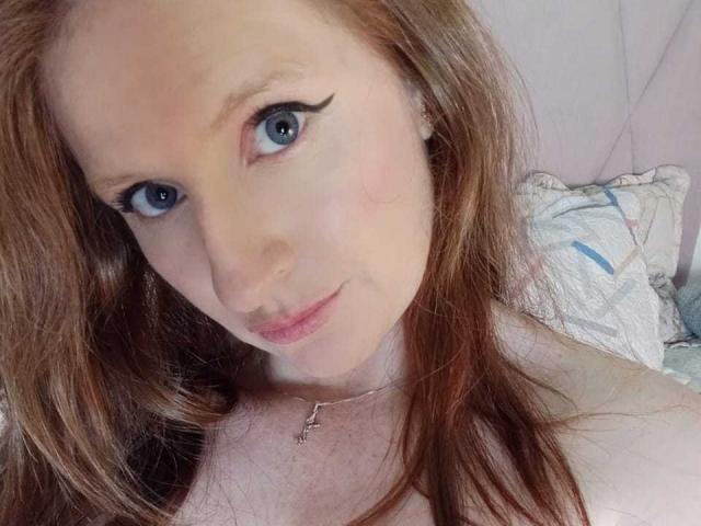 EnzumGinger has blue eyes, red hair, and a slight smile, wearing a necklace with a small cross, showing her bare shoulders. Her eyelashes are long and dark.