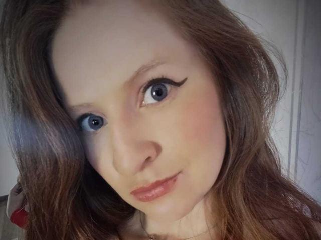 EnzumGinger has light blue eyes, thick brown wavy hair, and subtle makeup with black eyeliner. She's smiling slightly with pink lips. Her skin is fair and she's looking at camera.