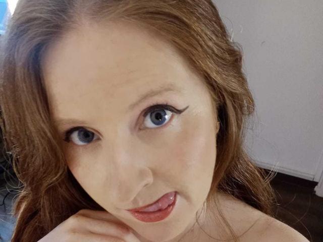 EnzumGinger has blue eyes, long red hair, and is sticking her tongue out with her hand near her chin. Her lips are slightly parted, showing a bit of her tongue.