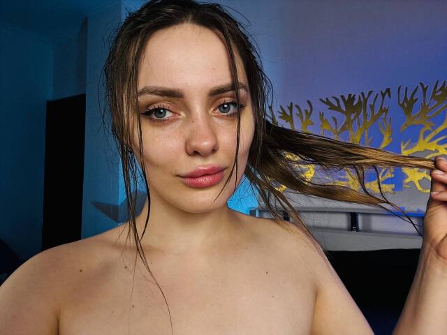 TenderAmberr with wet brown hair, holding a strand, has blue eyes, pink lips, and exposed bare shoulders, looking at the camera with a slight smile.